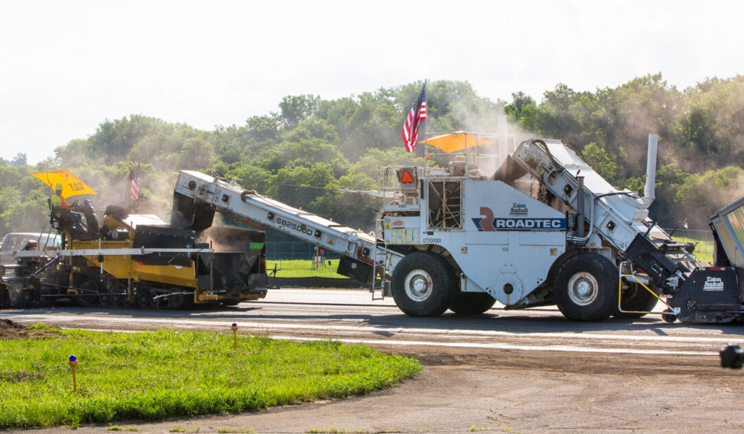 A photo of an Eaton Asphalt shuttle buggy working with a paver to pave the Lunken Airport runway in July 2019.
